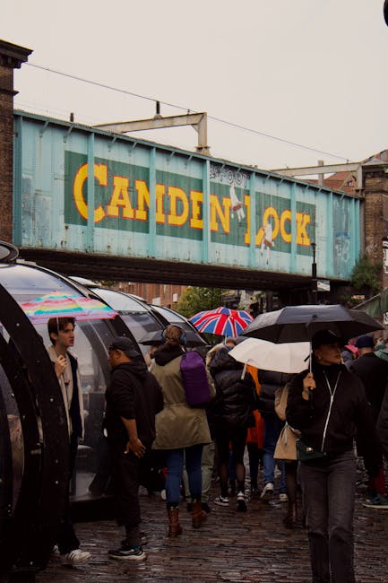 A group of people gathered on a cobbled street under a weathered blue metal railway bridge bearing the name 'CANDEN LOCK' in faded yellow letters, during a rainy day. Many of the pedestrians are holdi
