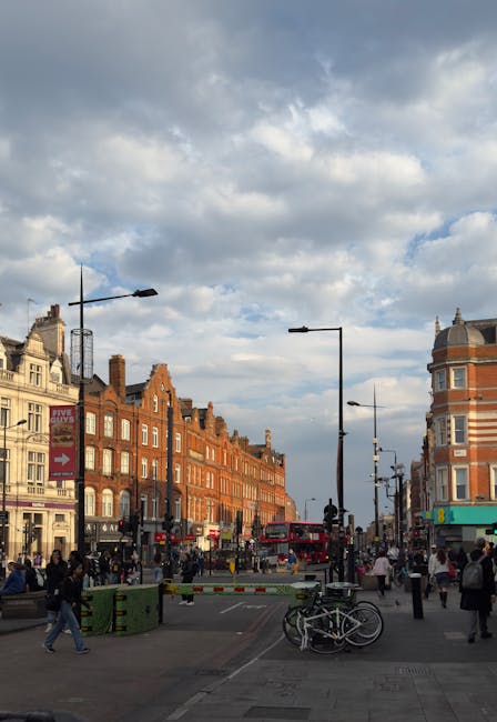 A busy street scene in Camden Town, NW1, showing a mix of brick and stone buildings lining the pavement, with pedestrians walking along the sidewalks and crossing the street. Several streetlights and 
