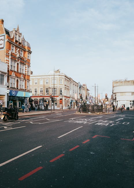 A busy street scene in Camden Town, NW1, showing a mix of brick and stone buildings lining the pavement, with pedestrians walking along the sidewalks and crossing the street. Several streetlights and 